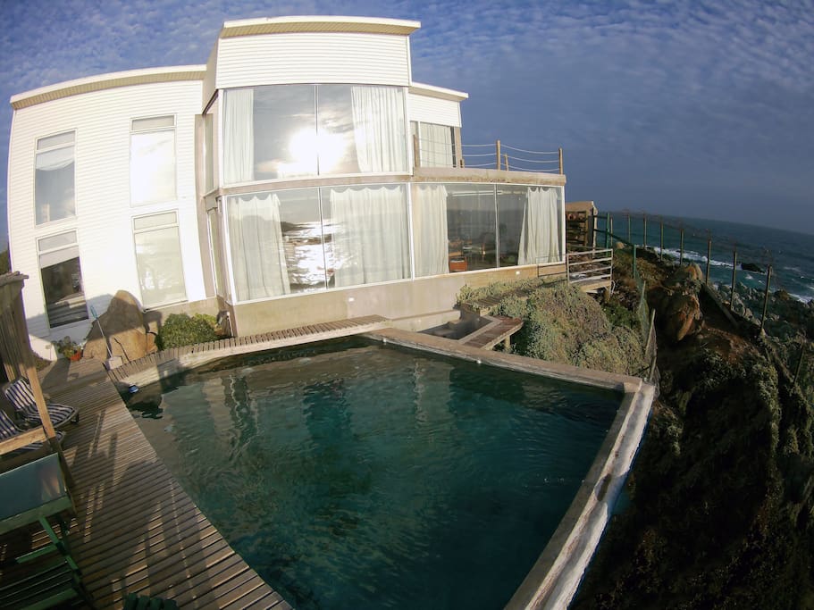 La casa Blanca es de 2 pisos, de 160 metros2, ubicada en primera línea frente a playa Isla Negra y con vista a Casa de Pablo Neruda, en condominio con guardia 24 horas y alarma de seguridad. Tiene 4 dormitorios, sala de estar, Living, Comedor, Cocina, bar, dormitorio principal con terraza, 3 baños, terraza exterior frente a piscina temperada a 27 grados (Excepto meses de mayo a agosto), jardines y amplio quincho. La casa está 100% equipada para disfrutar de unos agradables días en Isla Negra.