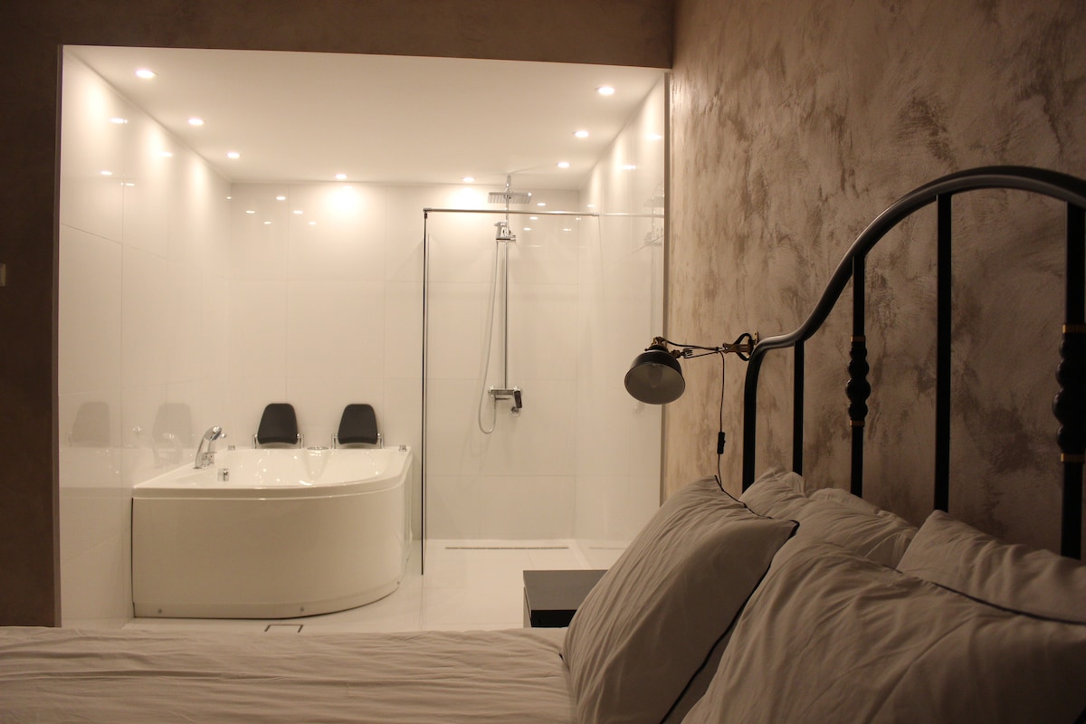 The open bedroom features a spacious bed with white linens and decorative pillows. A contemporary bathroom is visible, showcasing a sleek, modern designed jacuzzi tub, a glass-enclosed shower, and elegant lighting that enhances the clean aesthetic.