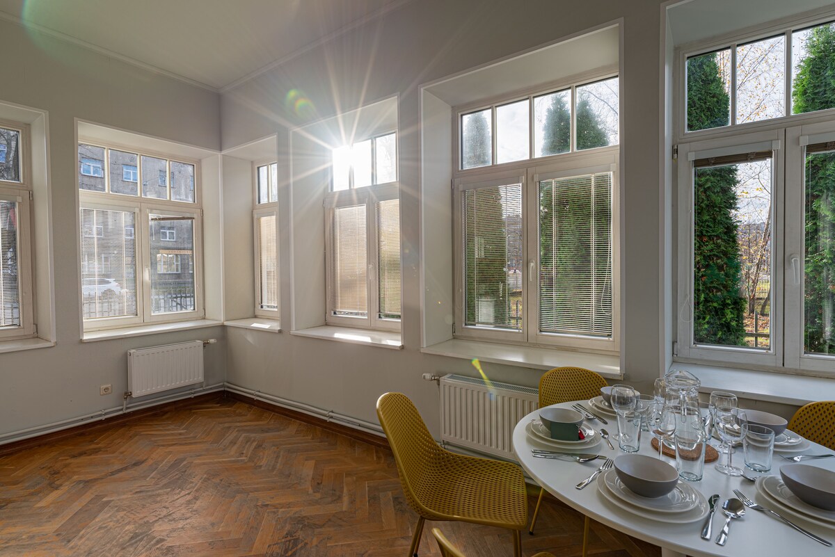 A dining area is illuminated by natural light streaming through multiple large windows. A round table is set with elegant dinnerware, while the rich wooden floor contrasts with the light-colored walls. Surrounding greenery can be seen through the windows, enhancing the space's openness.