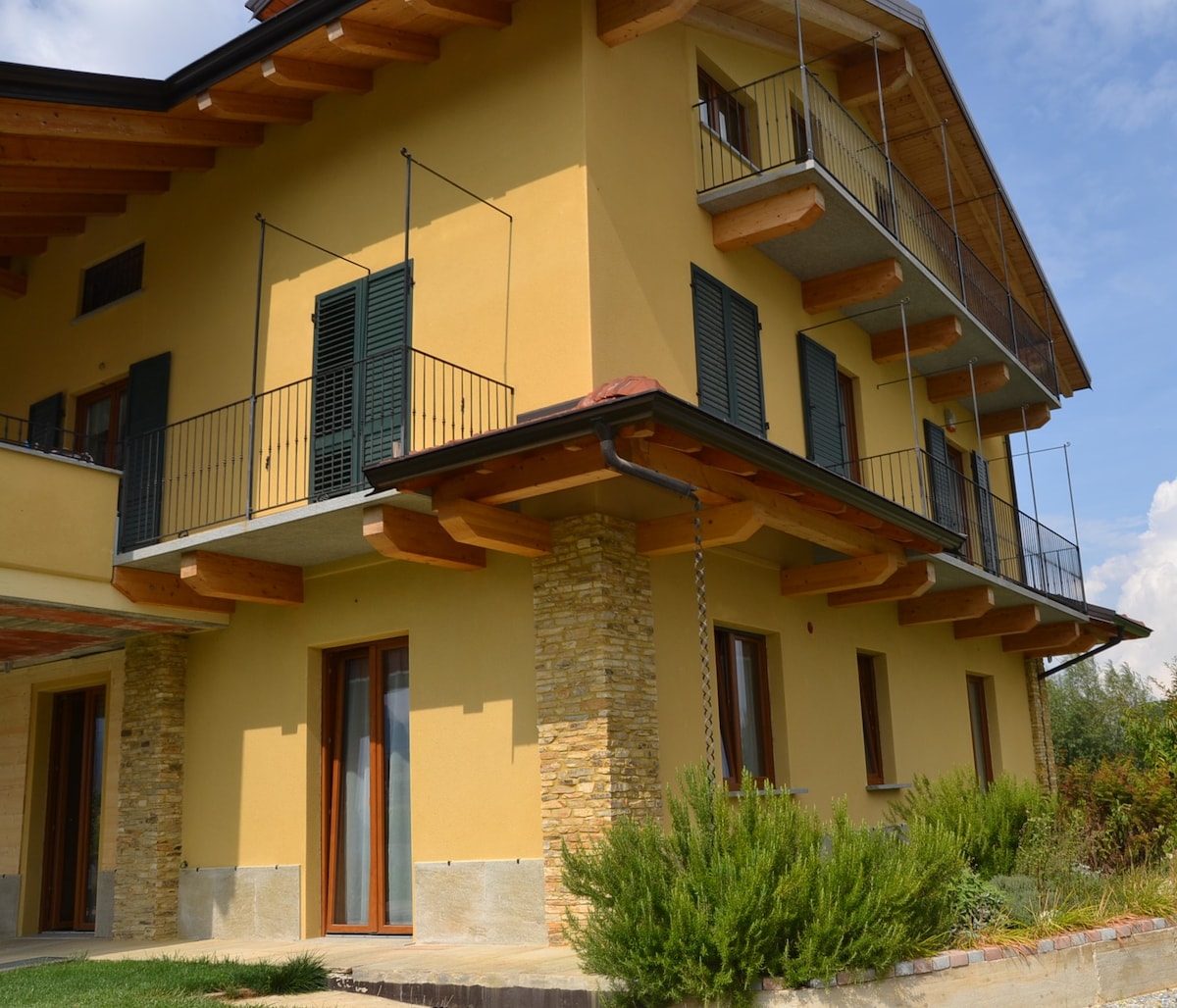 The exterior of the building features a warm yellow facade complemented by wooden balconies and stone accents. Large windows are framed by green shutters, and a landscaped area with greenery surrounds the entrance, enhancing the inviting appearance of the B&B.