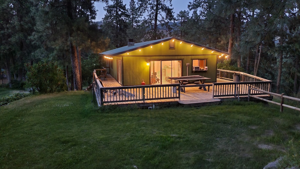 A remodeled house is surrounded by forest, featuring a wraparound deck illuminated by string lights. Large glass doors offer views into the interior space, while a picnic table is positioned on the deck, providing an inviting outdoor area against the natural backdrop.