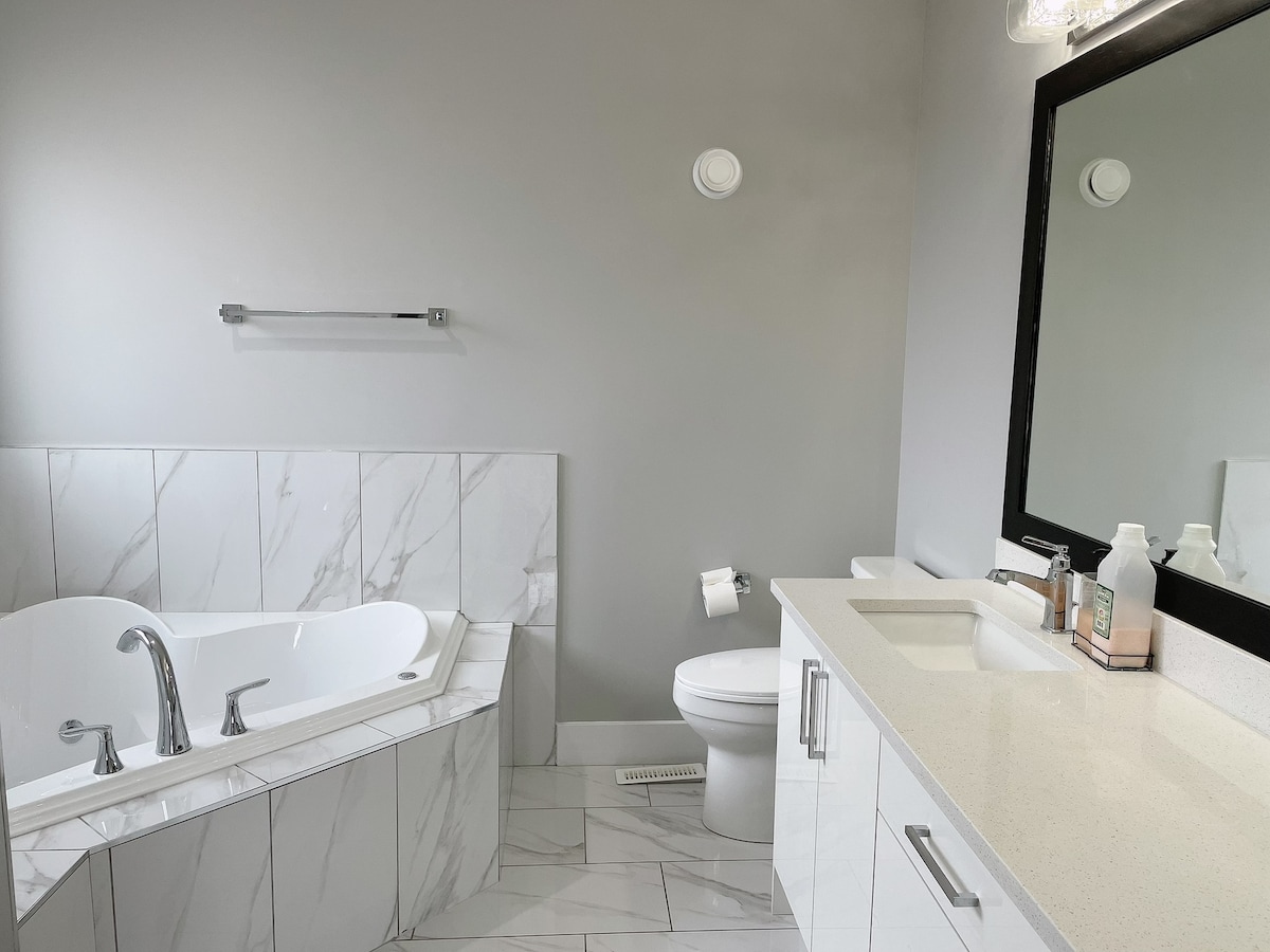 A modern bathroom features a freestanding tub positioned beside elegant marble tiles. A spacious, white vanity complements a large mirror, while a clean toilet is situated in the corner. Natural light reflects on the surfaces, enhancing the minimalist design.