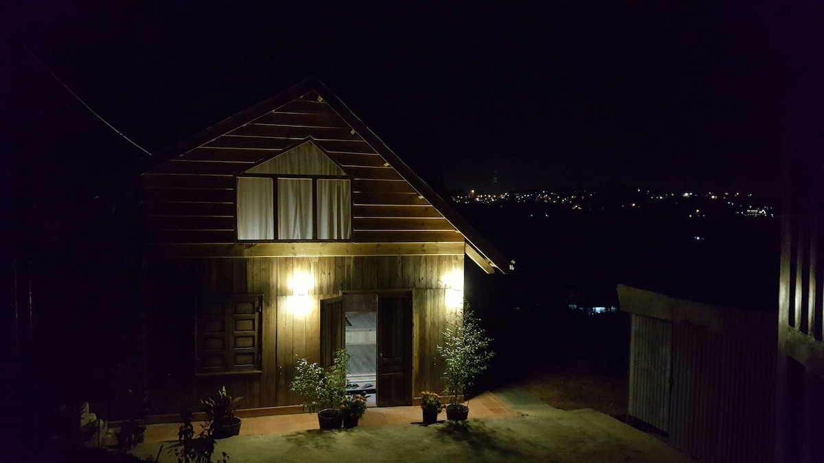 A wooden house is softly illuminated by exterior lights, highlighting the door and windows. Potted plants are placed near the entrance, while the night sky and distant city lights create a serene backdrop. The overall setting exudes a calm and inviting ambiance.