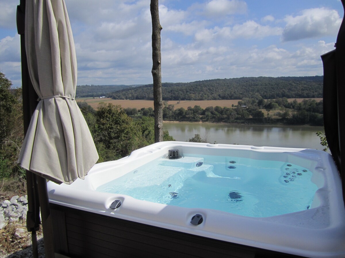 A private hot tub is situated on the deck, offering views of the Ohio River and vast rural landscapes. The water is clear, reflecting the sky, while surrounding trees provide a sense of serenity.