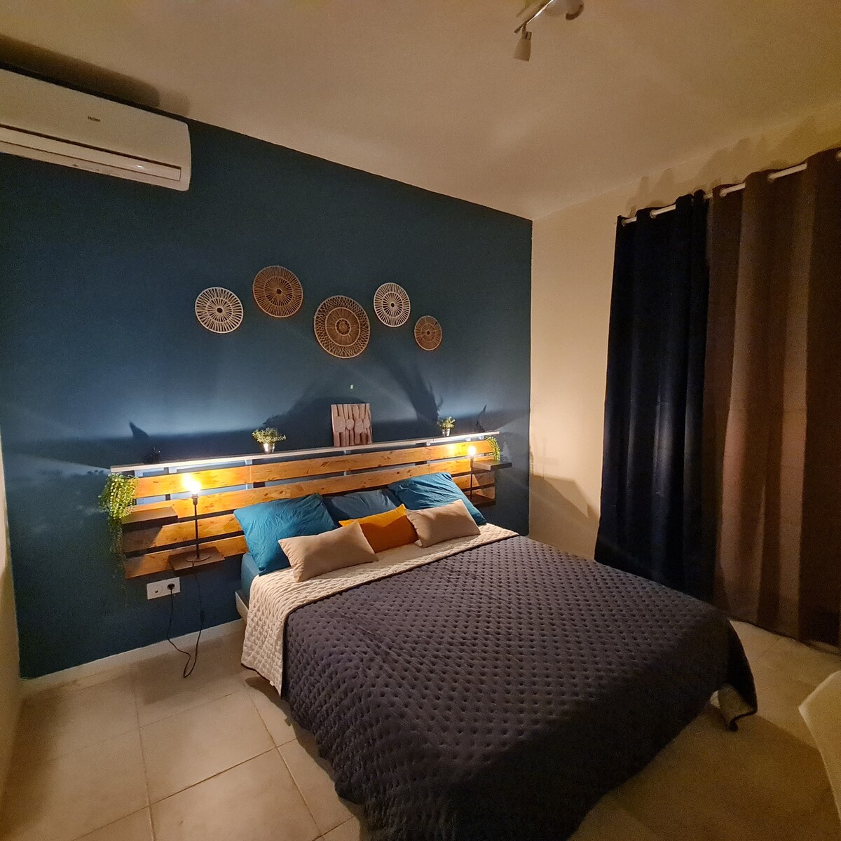 The bedroom features a spacious bed with a textured navy coverlet and decorative pillows. A wooden headboard is adorned with wall art and small plants, while ambient light creates a serene atmosphere. Dark curtains provide privacy, enhancing the room's tranquil feel.