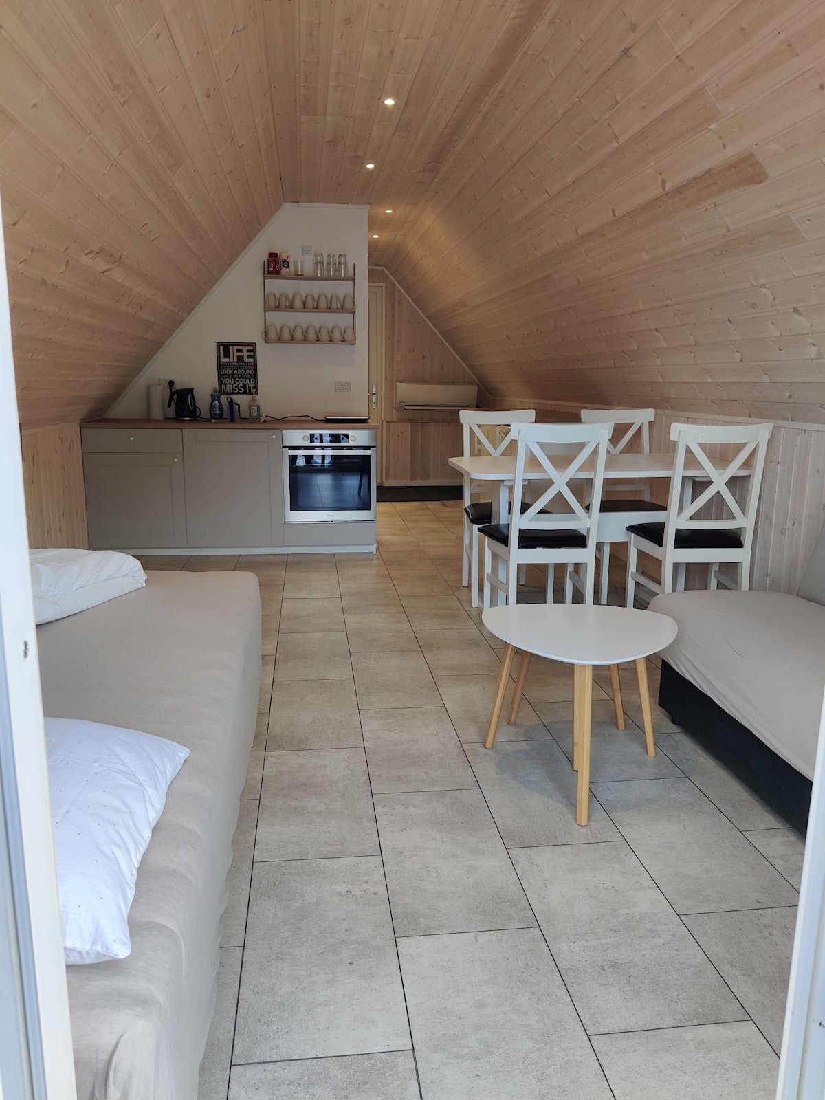 An interior space features a sloped wooden ceiling with soft lighting. A cozy seating area includes a beige sofa and a black couch, complemented by a light wood table. A dining area with white chairs and a small kitchen space is visible in the background.