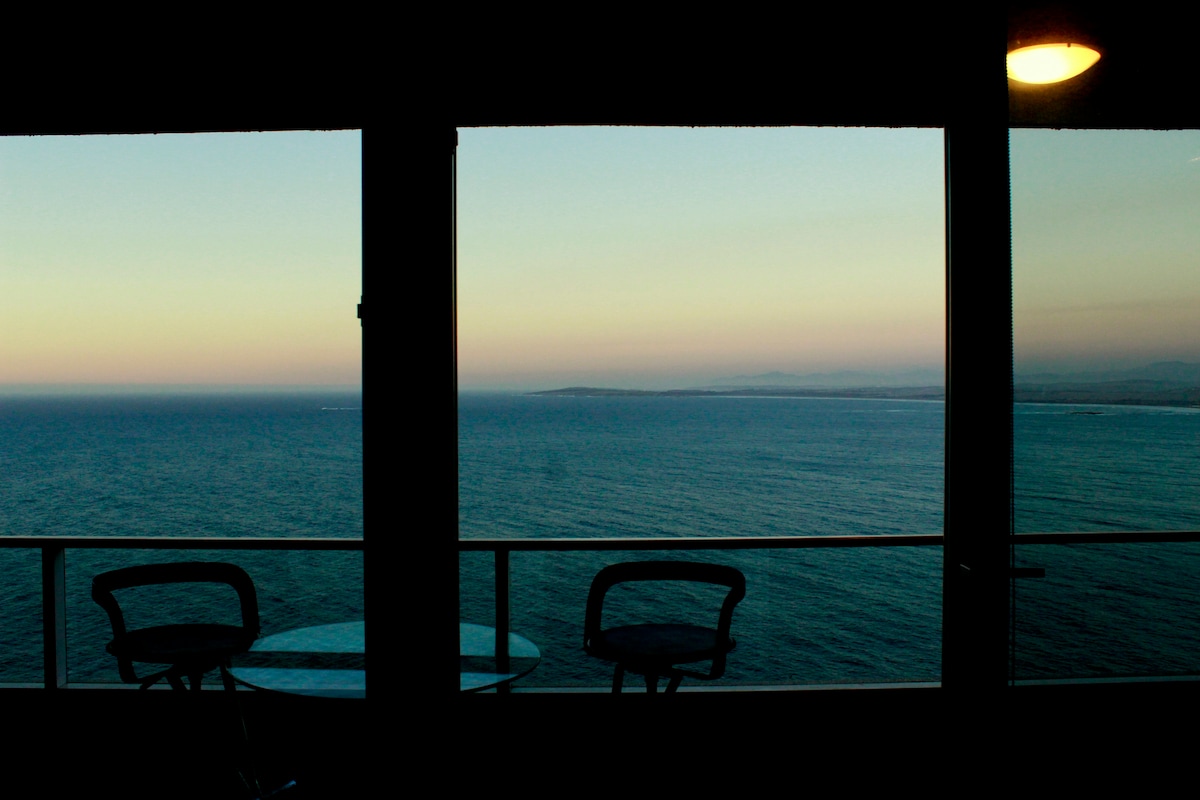 An expansive view of the ocean is framed by large windows, with a balcony featuring two chairs and a small table. The horizon displays soft colors of the sunset, reflecting the calmness of the sea. The room is bathed in soft natural light from the evening sky.