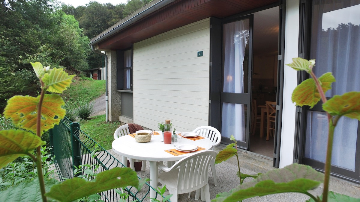 A shaded outdoor patio is furnished with a round table and four white chairs. Potted plants and tableware are arranged on the table. The entrance is visible through glass doors, leading to the interior space, which is surrounded by greenery.