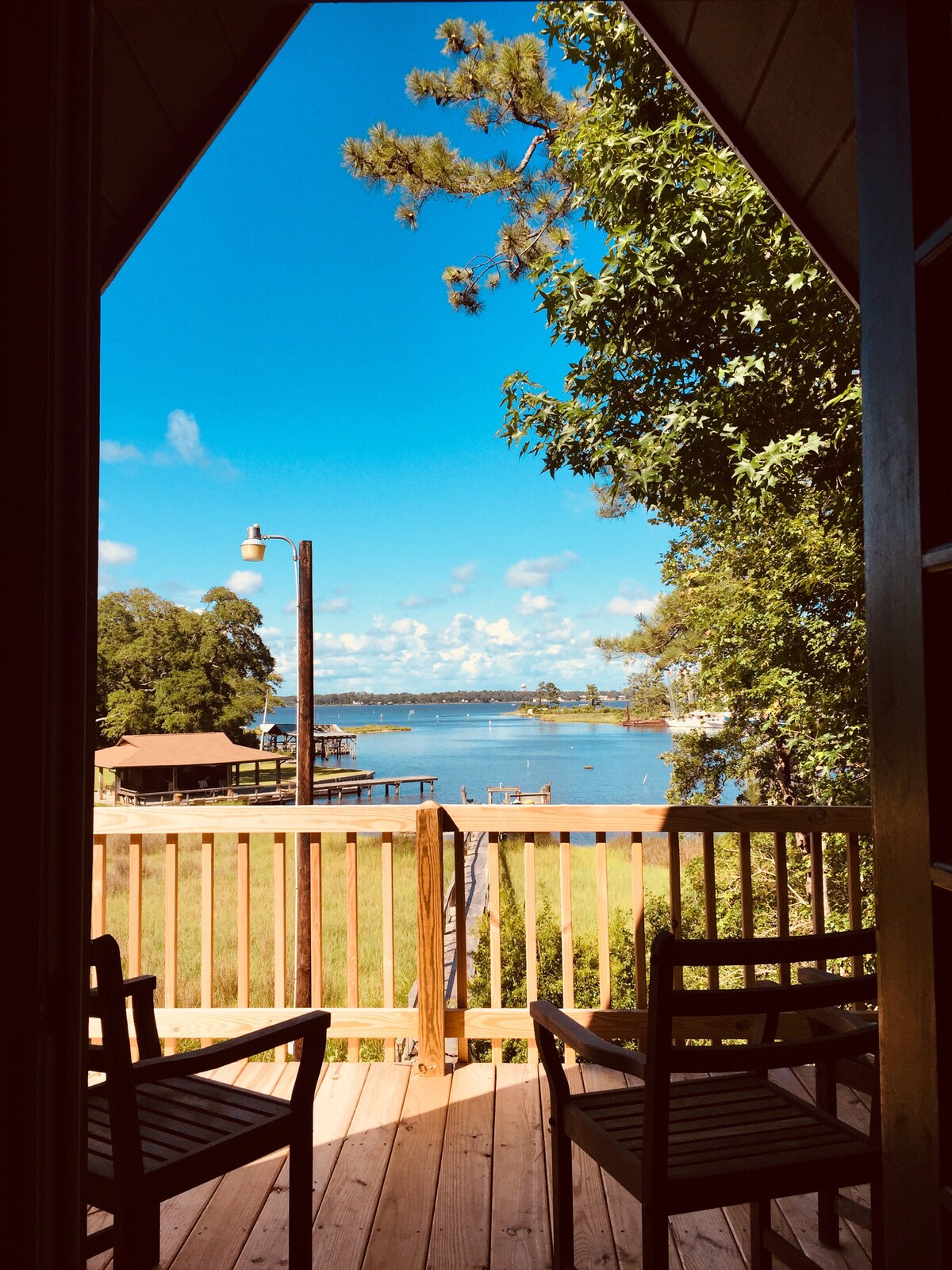 A private deck overlooks the serene waterfront, featuring two wooden chairs positioned for optimal views. The sky is bright blue with scattered clouds, and the water reflects the natural beauty of the surrounding landscape, inviting guests to relax in a tranquil setting.