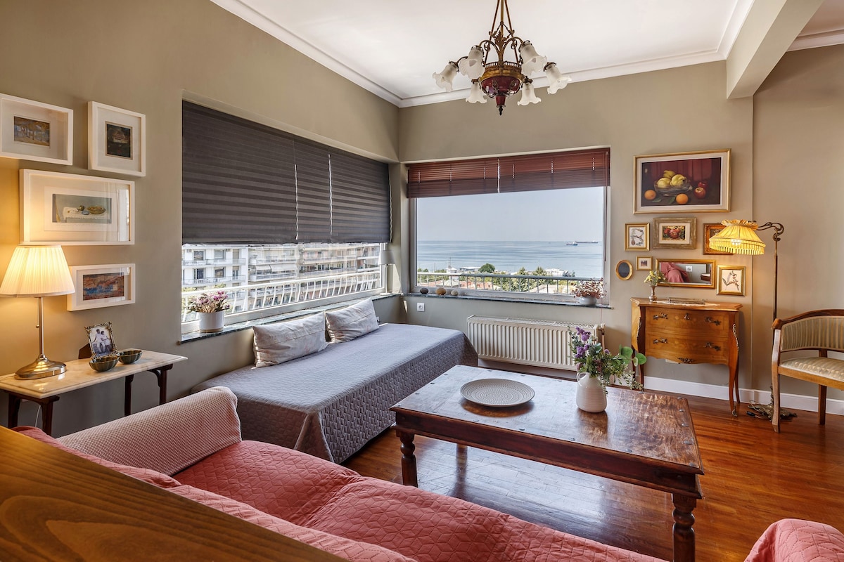 A bright living space features a large window with unobstructed sea views. Comfortable seating includes a plush sofa and a simple lounge chair. A wooden table, adorned with fresh flowers, sits at center, while framed artwork and lamps add a touch of warmth to the room.