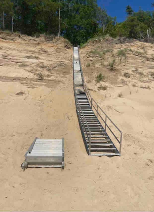 A long metal ramp is positioned on a sandy incline, leading towards a beach area. The ramp features a slight rise, facilitating access from the sandy ground to higher terrain. Green trees line the top of the incline under a clear blue sky.