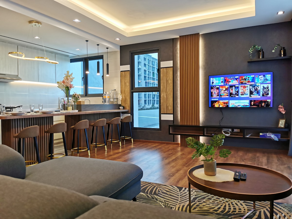 A modern living space features a comfortable seating area with a dark sectional sofa and a coffee table. A wall-mounted 55" Smart TV displays a variety of media options. The kitchen island, equipped with bar stools, is complemented by stylish pendant lighting and large windows offering city views.