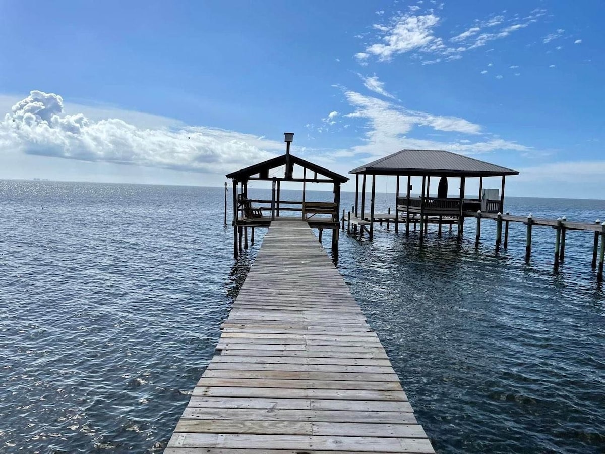 A wooden pier extends into Mobile Bay, flanked by two structures. The left structure offers seating, while the right features a roof. The calm water reflects the blue sky, with soft clouds scattered above, creating a serene atmosphere for relaxation and observation.