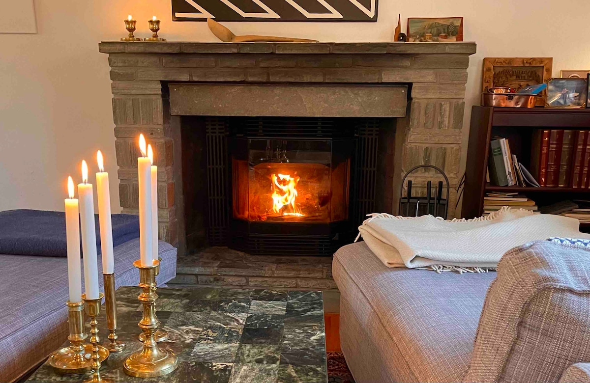 A cozy living area features a stone fireplace with a warm, flickering fire. Candles on a marble table provide soft lighting, while a blanket and a pillow are placed on the sofa, inviting relaxation in front of the hearth.