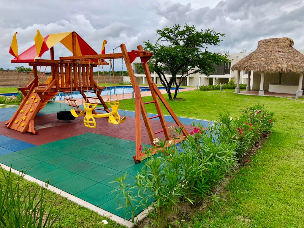 A colorful playground set features swings and a slide, positioned on a padded play surface. Surrounding greenery adds a refreshing touch, while a shaded palapa structure and a pool area are visible in the background, creating a recreational space for families.