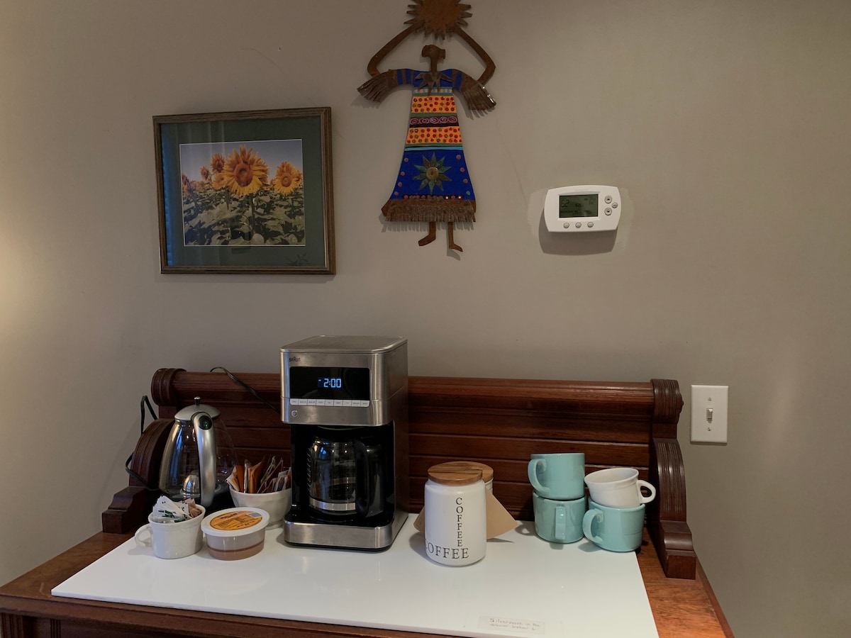 A coffee station is displayed with a coffee maker, kettle, and various mugs arranged neatly. A small bowl holds sugar packets, while a decorative wall art piece features a figure with vibrant colors. A thermostat is mounted on the wall above the setup.