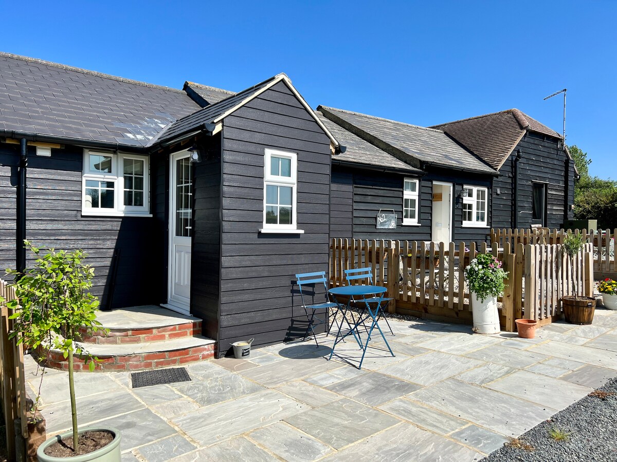 A charming exterior of a black wooden cottage is showcased under a clear blue sky. A welcoming entrance leads to neatly arranged outdoor seating with two small chairs and a table, surrounded by potted plants and a wooden fence, creating a serene outdoor space.