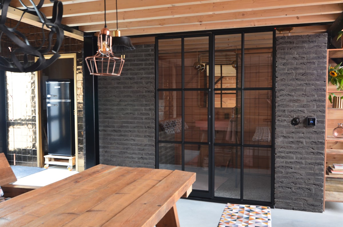 A modern loft space is presented with an industrial design, featuring a black-framed glass partition that separates the living area from the adjoining room. A rustic wooden dining table is positioned in the foreground, complemented by stylish light fixtures and exposed wooden beams overhead.