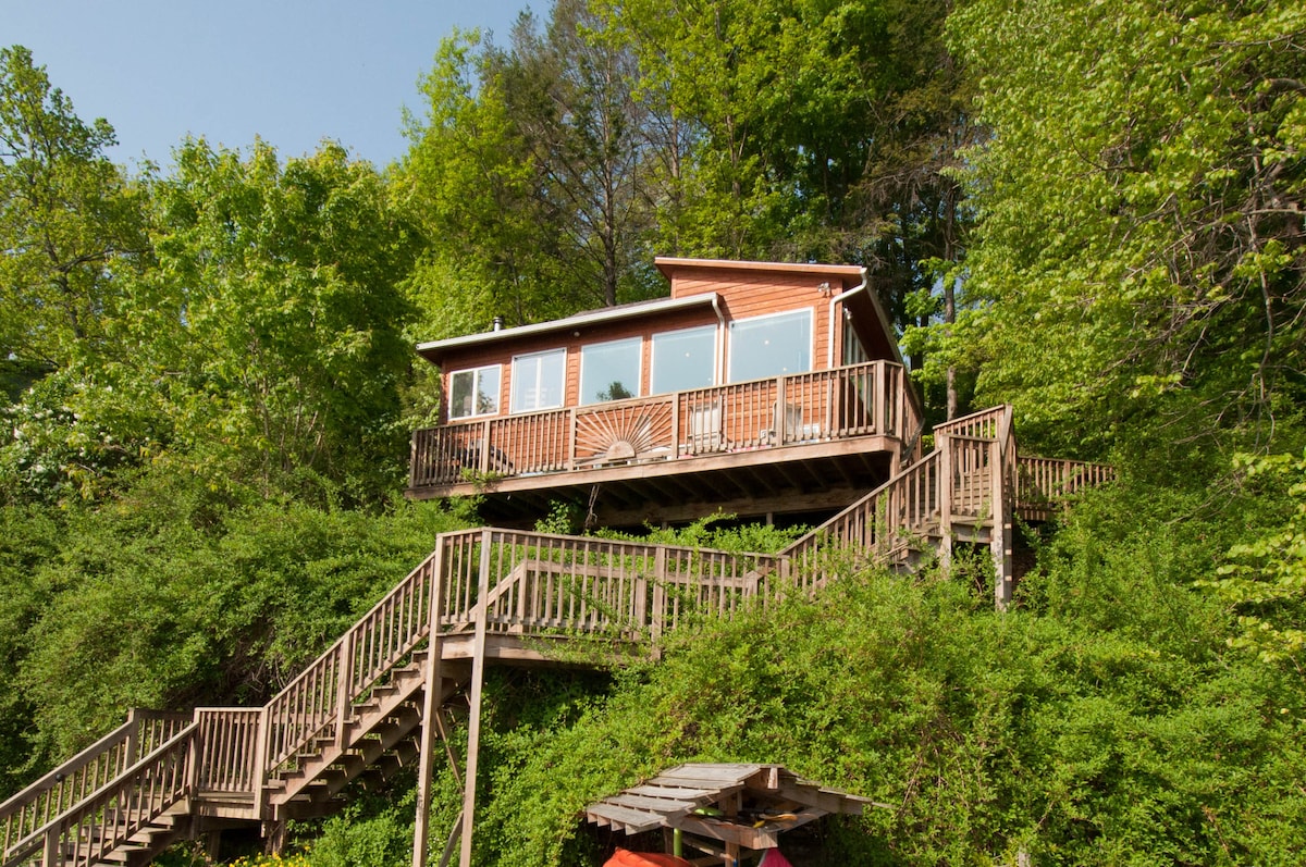 A lakeside home is nestled among lush greenery, elevated on a hillside. A series of wooden stairs lead from the ground to the spacious deck, which features large windows overlooking the lake. The surrounding trees provide natural shade and a sense of privacy.