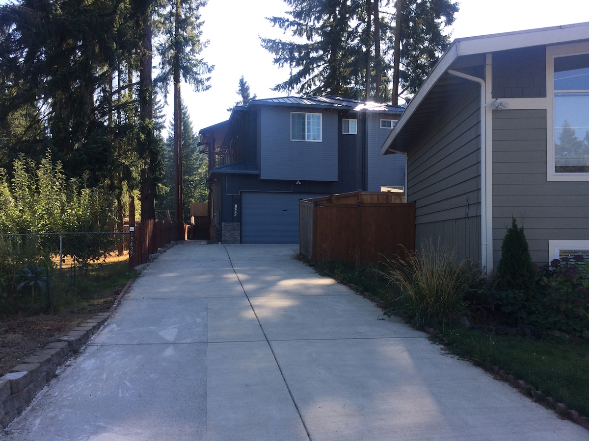 A paved driveway leads to a multi-level house accented by large windows, surrounded by tall evergreen trees. A neighboring building is visible on the right, and wooden fences provide a sense of privacy. The spacious outdoor area is well-maintained.
