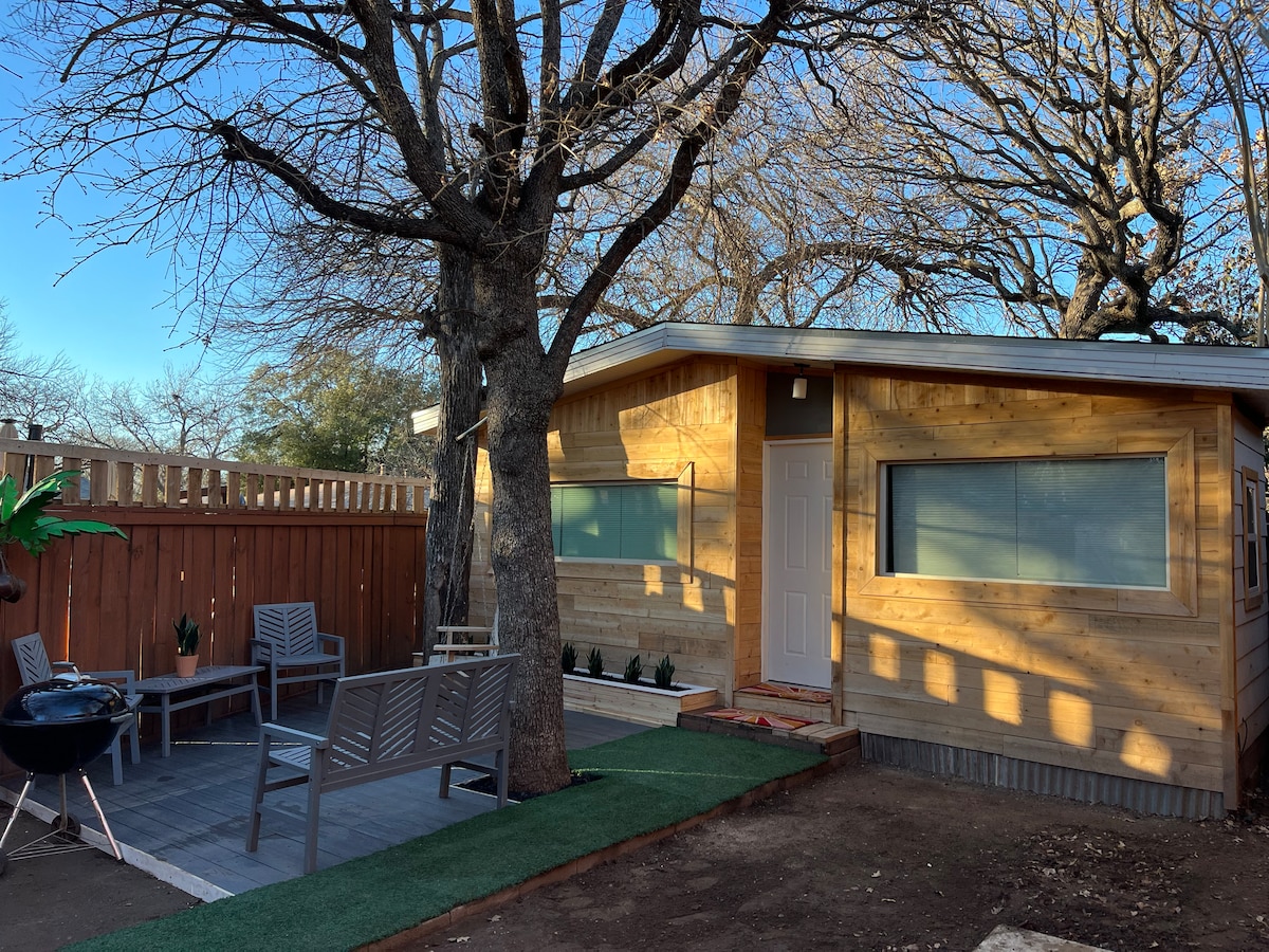 A single-story wooden building features large windows that allow natural light to fill the space. Outside, a small patio area is furnished with seating and a grill, surrounded by neatly arranged greenery and a well-maintained lawn.