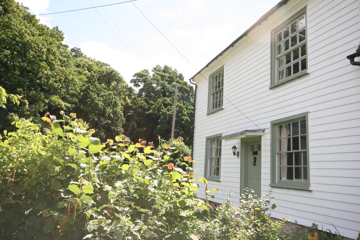 The exterior of the farmhouse showcases a charming white facade with green trim. Quiet gardens filled with lush greenery and blooming flowers surround the building, providing a serene and inviting atmosphere. Large windows allow natural light to illuminate the interior spaces.