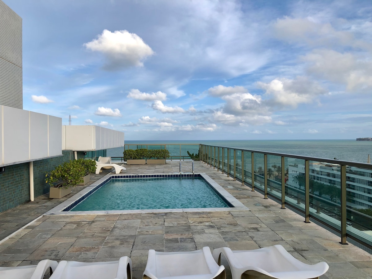 A rooftop terrace features a swimming pool surrounded by smooth stone tiles. Lounge chairs are positioned along the edge, providing views of the ocean and sky. The serene atmosphere is enhanced by the presence of greenery in planters along the perimeter.