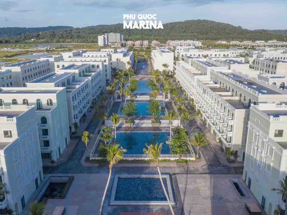 Aerial view captures a spacious resort area featuring multiple swimming pools lined with palm trees. The tranquil blue waters of the pools contrast with the white architecture of nearby buildings, nestled against a backdrop of lush green hills.