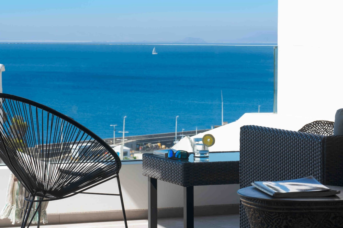 A terrace area features a stylish wicker table with a glass top, accompanied by a comfortable chair. The sea stretches out in the background, showcasing different shades of blue, while a distant sailboat glides across the horizon.