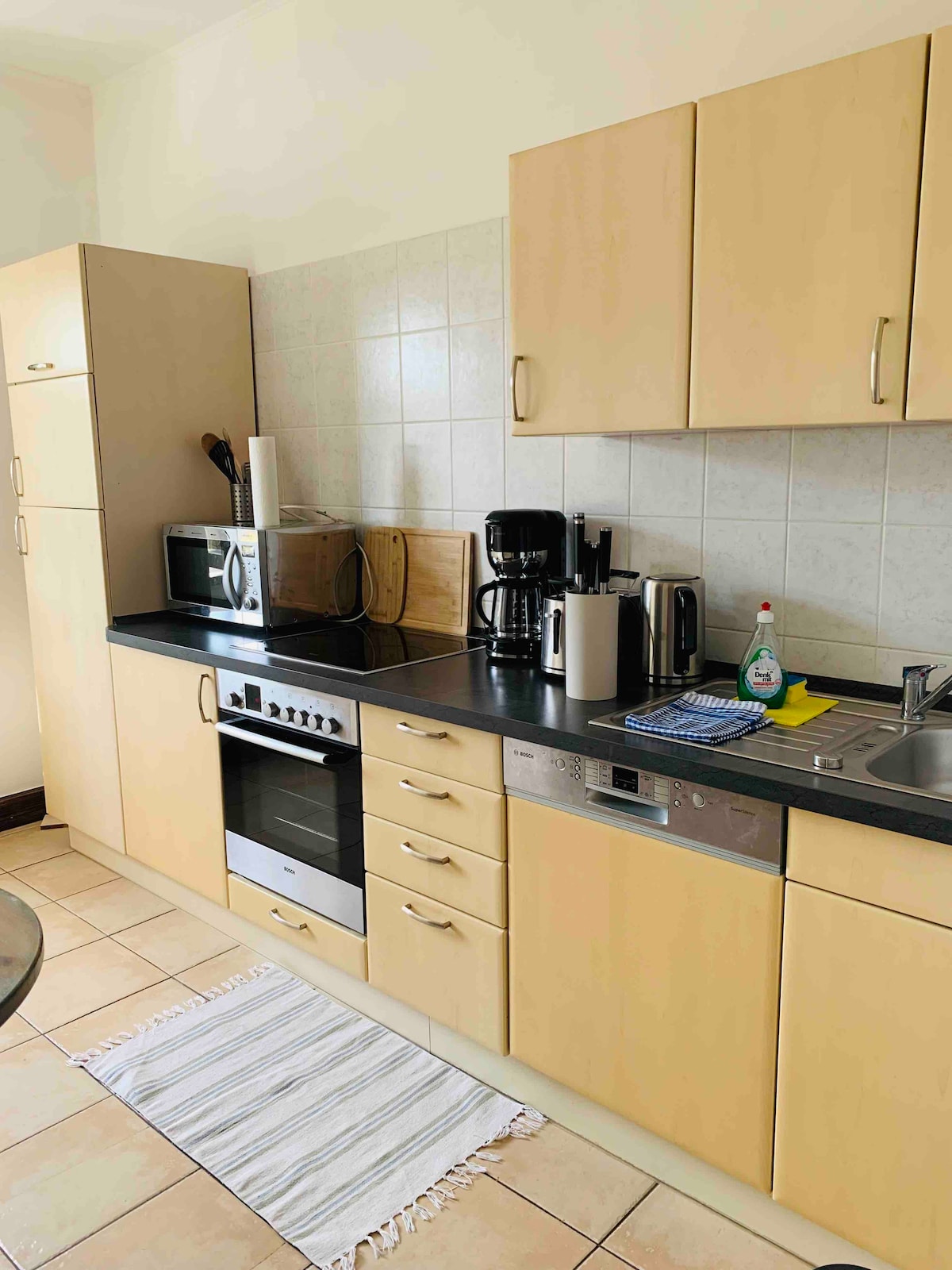 The kitchen features light-colored cabinetry and a dark countertop. Equipped with a stovetop, oven, microwave, coffee maker, and dishwasher, the space is designed for functionality. A small dish drying rack and cleaning supplies are present, along with a light-colored rug on the tiled floor.