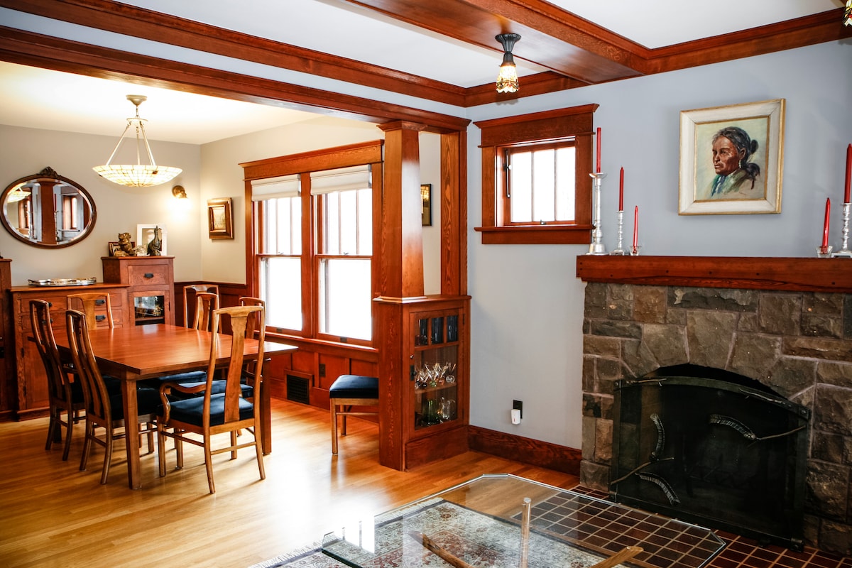 A dining area is presented with a wooden table surrounded by matching chairs. The room features crafted wood paneling and large windows that allow natural light to fill the space. A stone fireplace adds a cozy element, complemented by decorative items on the mantel.