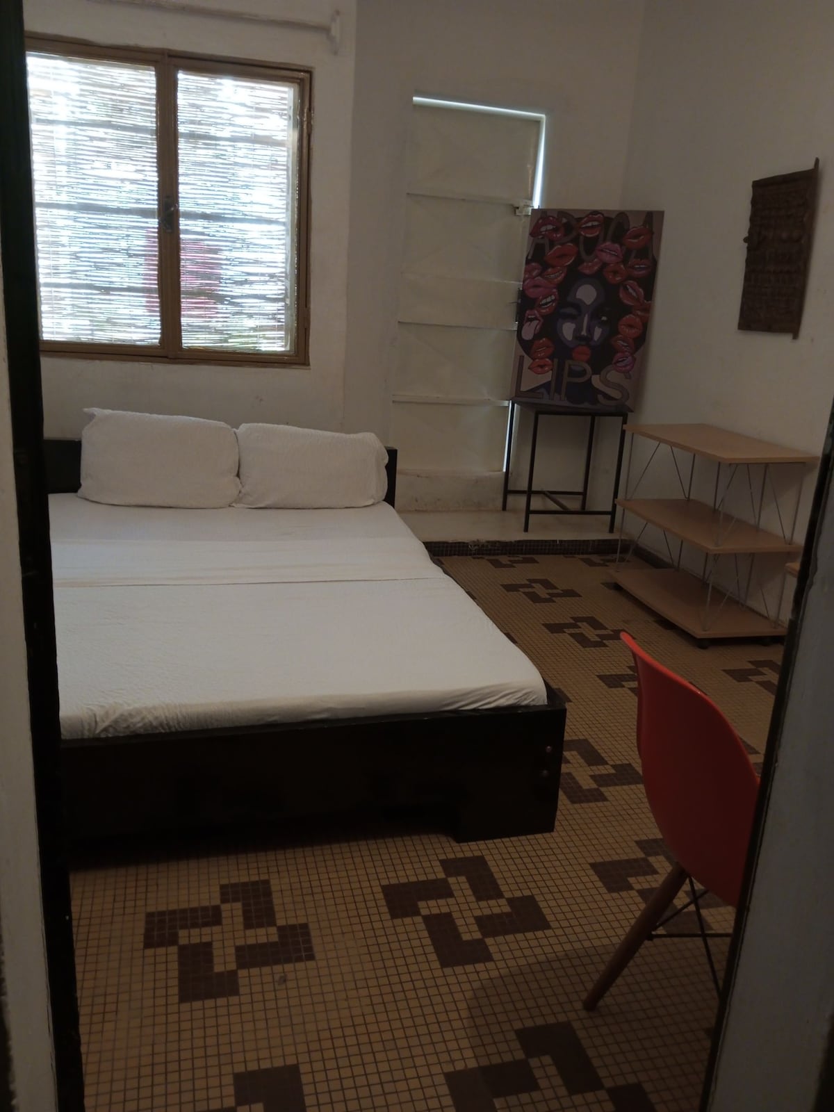 A simple bedroom features a spacious bed with white bedding, positioned against a light-colored wall. A window allows natural light into the room, complemented by a piece of artwork on display. A shelving unit and a red chair provide functional furnishings.