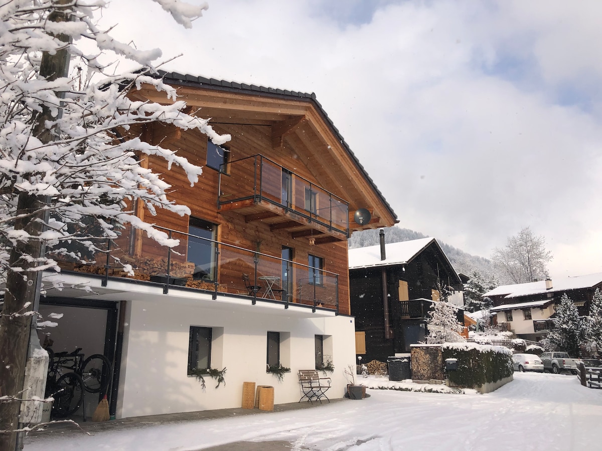 A charming wooden building stands amidst winter snow, featuring large glass windows and a balcony that overlooks the landscape. Adjacent structures display a traditional alpine style, while a paved area with parked bicycles and snow-dusted trees enhance the serene outdoor environment.