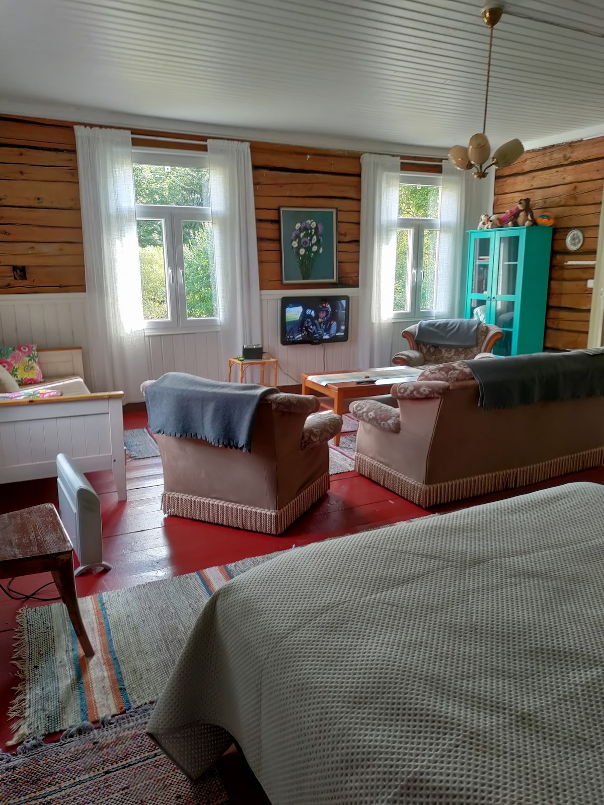 A cozy living area features two armchairs draped with light blankets and a comfortable sofa. A wooden coffee table stands between the seating, while a television is mounted on the wall. Natural light fills the room through two windows, and a colorful rug adds warmth.