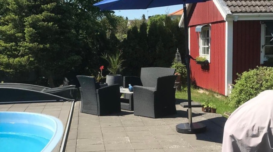 An outdoor patio area is depicted, featuring a circular pool and a small table surrounded by four dark wicker chairs. A large umbrella offers shade, while lush greenery surrounds a red cottage in the background.