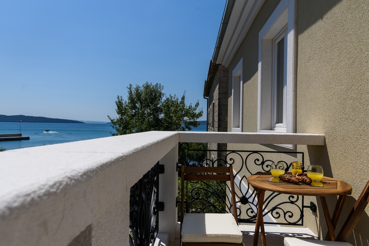 A balcony offers a clear view of the sea, with a small round table set for two. A pair of wooden chairs are placed beside the table, which is adorned with a fruit bowl and pastries. The railing features decorative ironwork, enhancing the outdoor space.