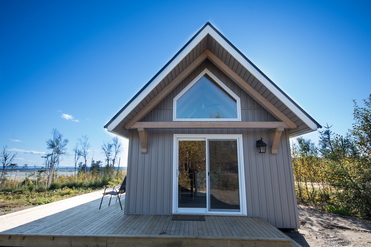A charming glamping unit is depicted with a sloped roof and large glass doors. A spacious wooden deck extends in front, offering a welcoming space. Two outdoor chairs are positioned by the entrance, surrounded by natural greenery that frames the scene.
