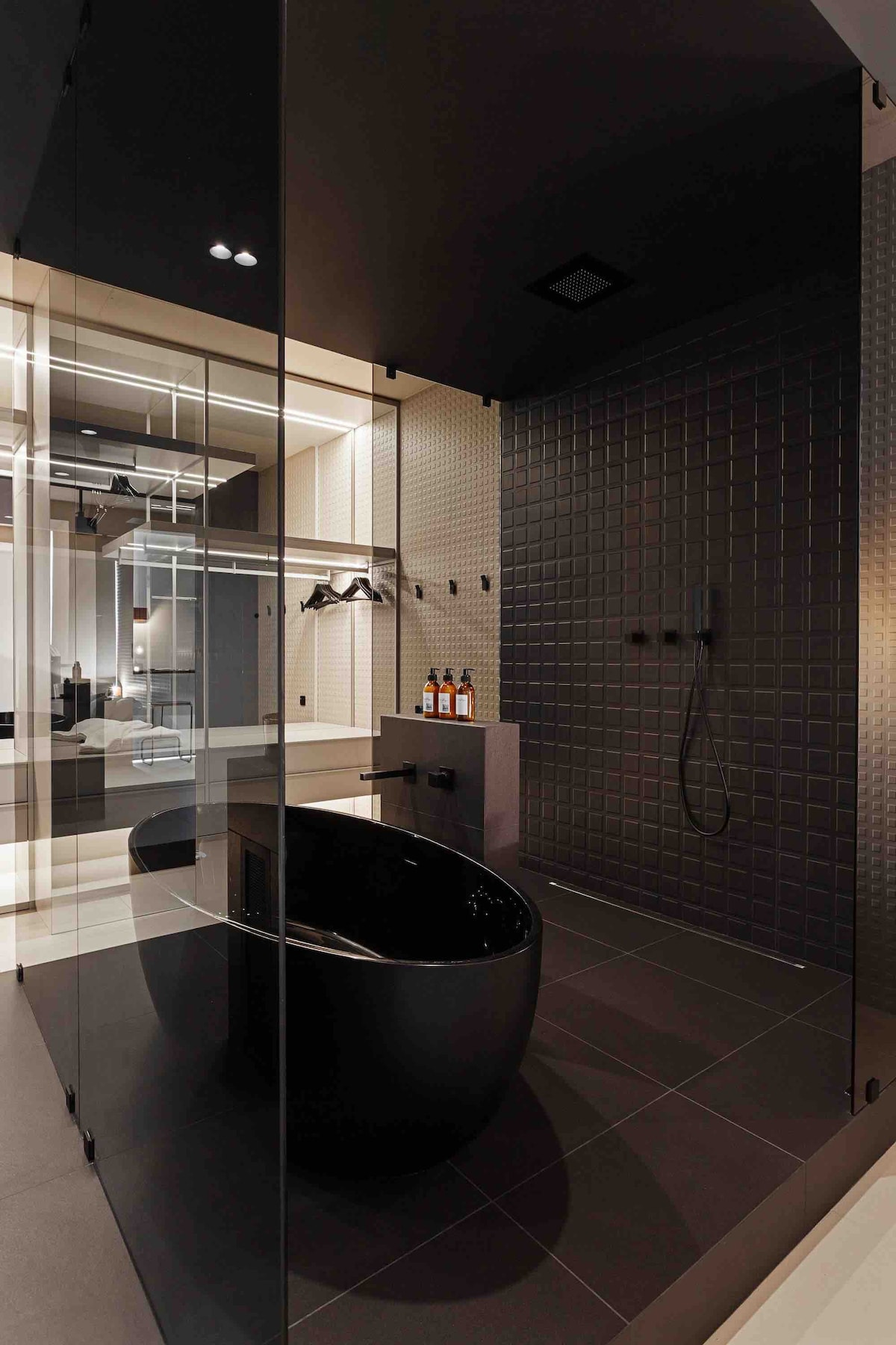 A modern bathroom is showcased with a sleek black freestanding tub and a spacious glass-enclosed shower. The walls are accented with textured tiles, offering a sophisticated appearance. Neutral lighting highlights the clean lines and minimalistic design of the fixtures and decor.