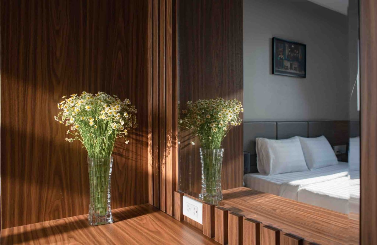 A simple yet elegant wooden vanity is shown, featuring a clear glass vase with fresh flowers. Reflections in the mirror display soft lighting and a neatly made spacious bed in the background, creating a serene atmosphere.