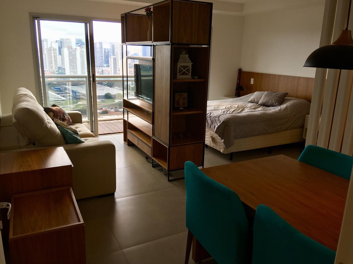 A well-furnished living space is displayed, featuring a spacious bed and a cozy seating area. Large windows allow natural light to illuminate the room and provide a view of the city. A wooden table with colorful chairs complements the modern design.