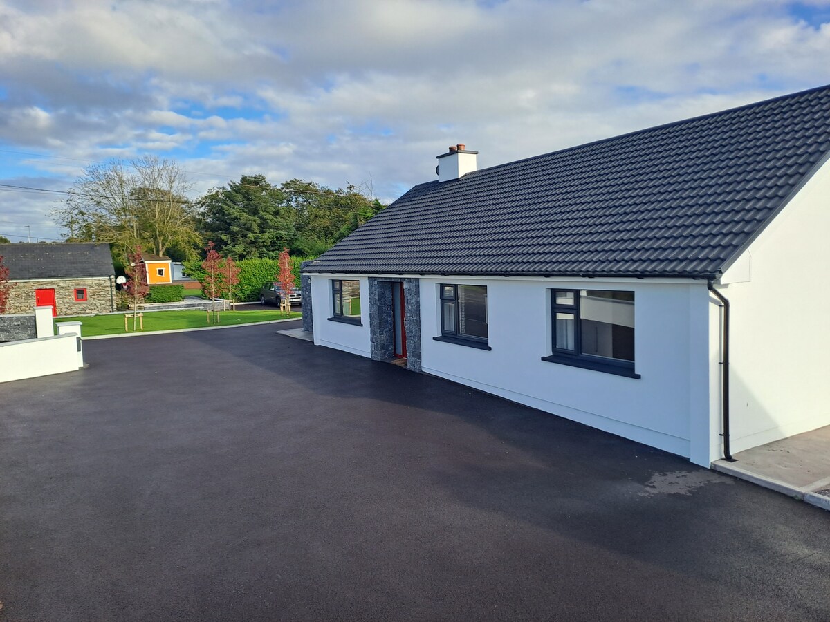 A renovated bungalow is positioned along a freshly paved path, featuring a smooth black driveway. Its white exterior contrasts with a slate-colored roof. Surrounding greenery and scattered trees create a pleasant setting. Calm skies are visible, enhancing the tranquil vibe of the property.