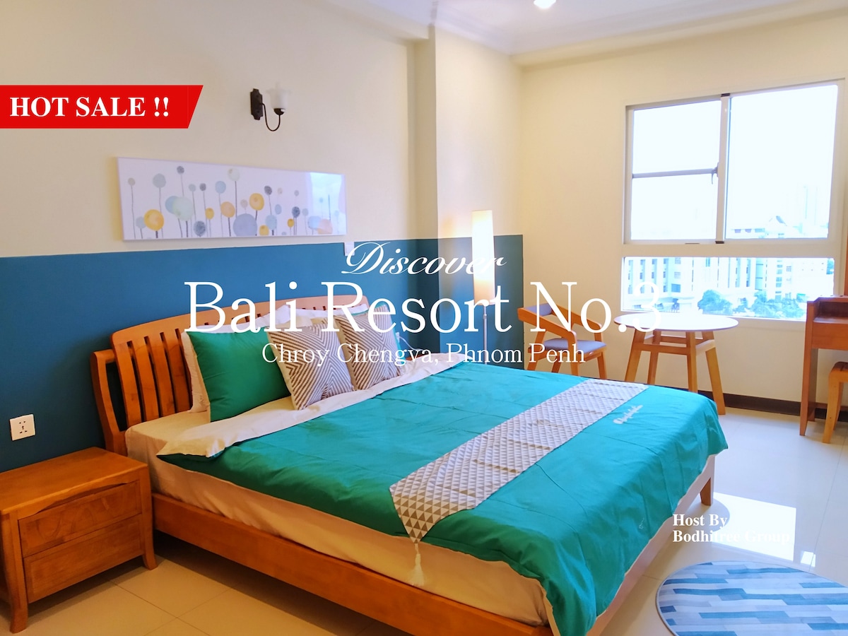 A well-furnished sleeping area features a spacious bed with vibrant bedding. A decorative wall art piece adds a touch of color. Natural light enters through a large window beside a wooden dining table and chairs, creating a cozy atmosphere.