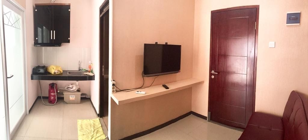 A compact living space features a small kitchenette with dark cabinetry and a washing area. A mounted television is positioned on a light-colored wall, complemented by a brown sofa across. Soft lighting enhances the simple and functional layout.