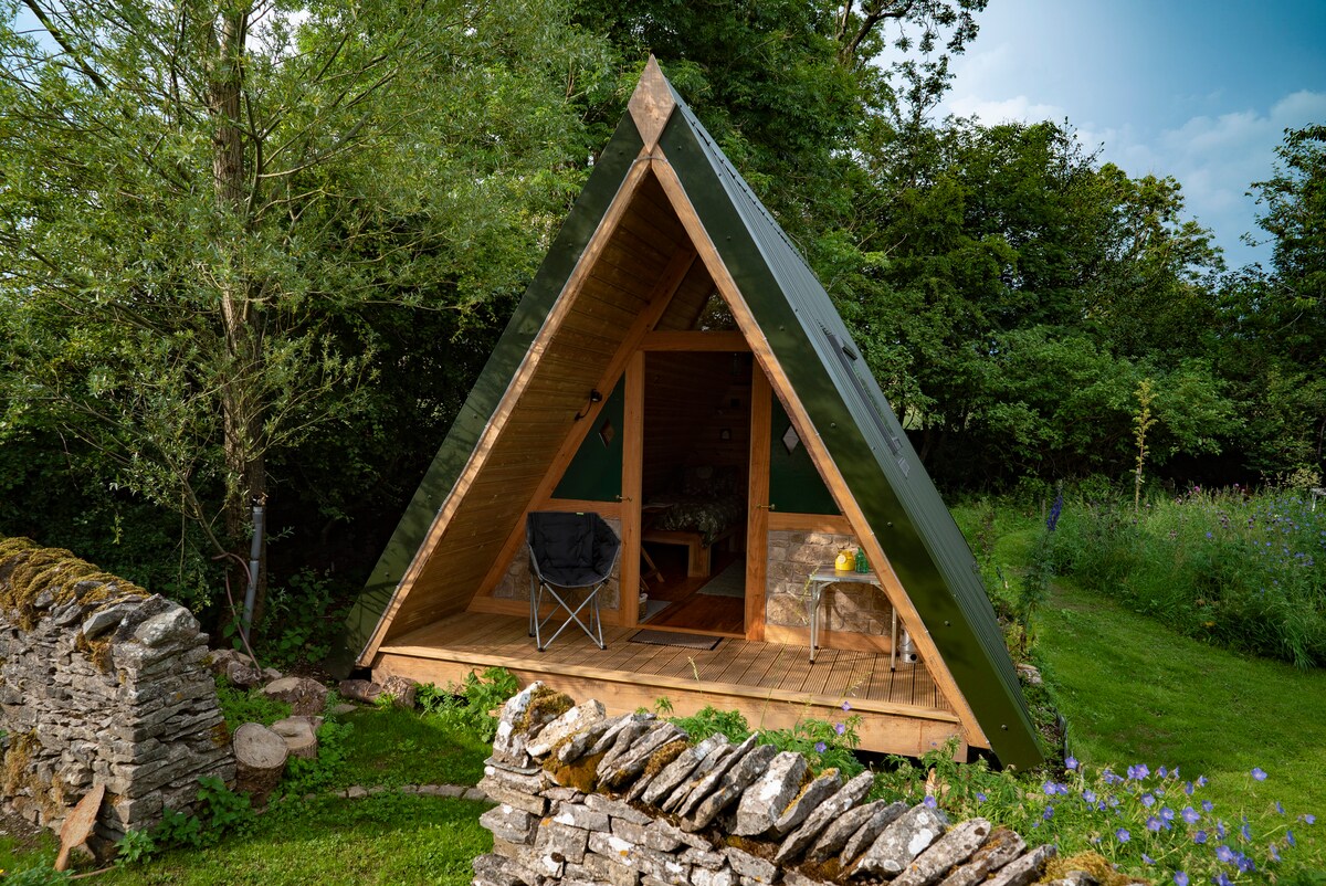 A wooden cabin with a green tin roof is set within a lush garden, framed by trees. The structure features a spacious deck area with a camping chair and a stone wall in the foreground, enhancing the natural surroundings.