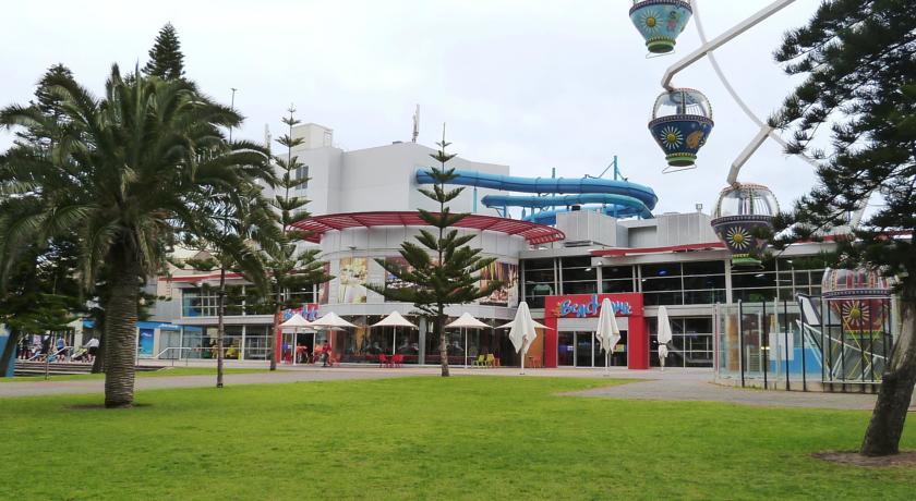 The image displays a modern beachfront complex with a slide and gondolas in the background. Palm trees line the foreground, framing a grassy area that leads to the building's entrance. Outdoor seating can be seen under large umbrellas, inviting guests to relax.