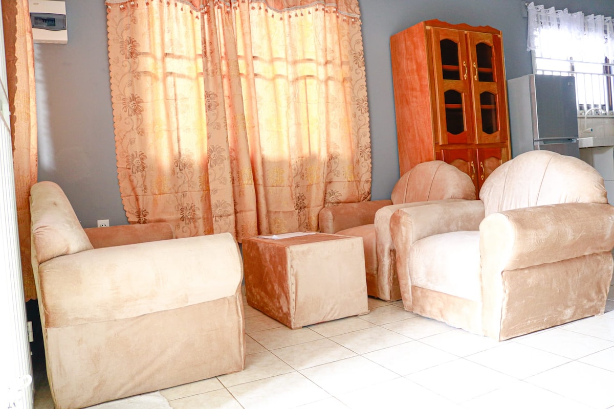 A comfortable seating area features two plush armchairs and a matching sofa, arranged around a simple coffee table. Light filters through sheer curtains, illuminating the soft beige upholstery. A wooden cabinet is positioned against the wall, providing additional storage space.