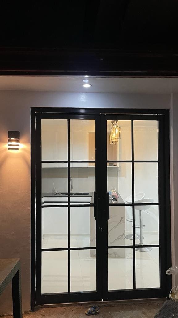 A modern glass door opens to reveal a well-lit kitchen area featuring sleek cabinetry and a minimalist design. Soft lighting highlights the space, while a bar stool can be seen near the counter, inviting a cozy dining experience.
