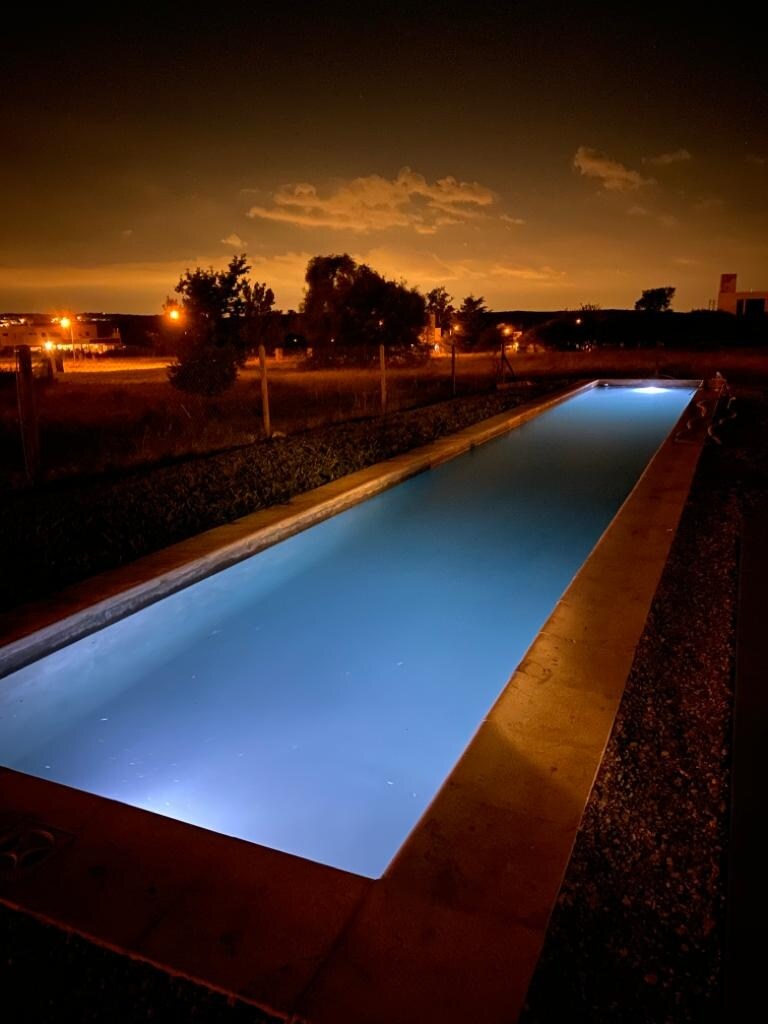 An illuminated swimming channel is visible at night, with soft turquoise lighting reflecting in the water. Surrounding greenery is suggested by silhouettes, while distant city lights are faintly discernible against a dark sky.