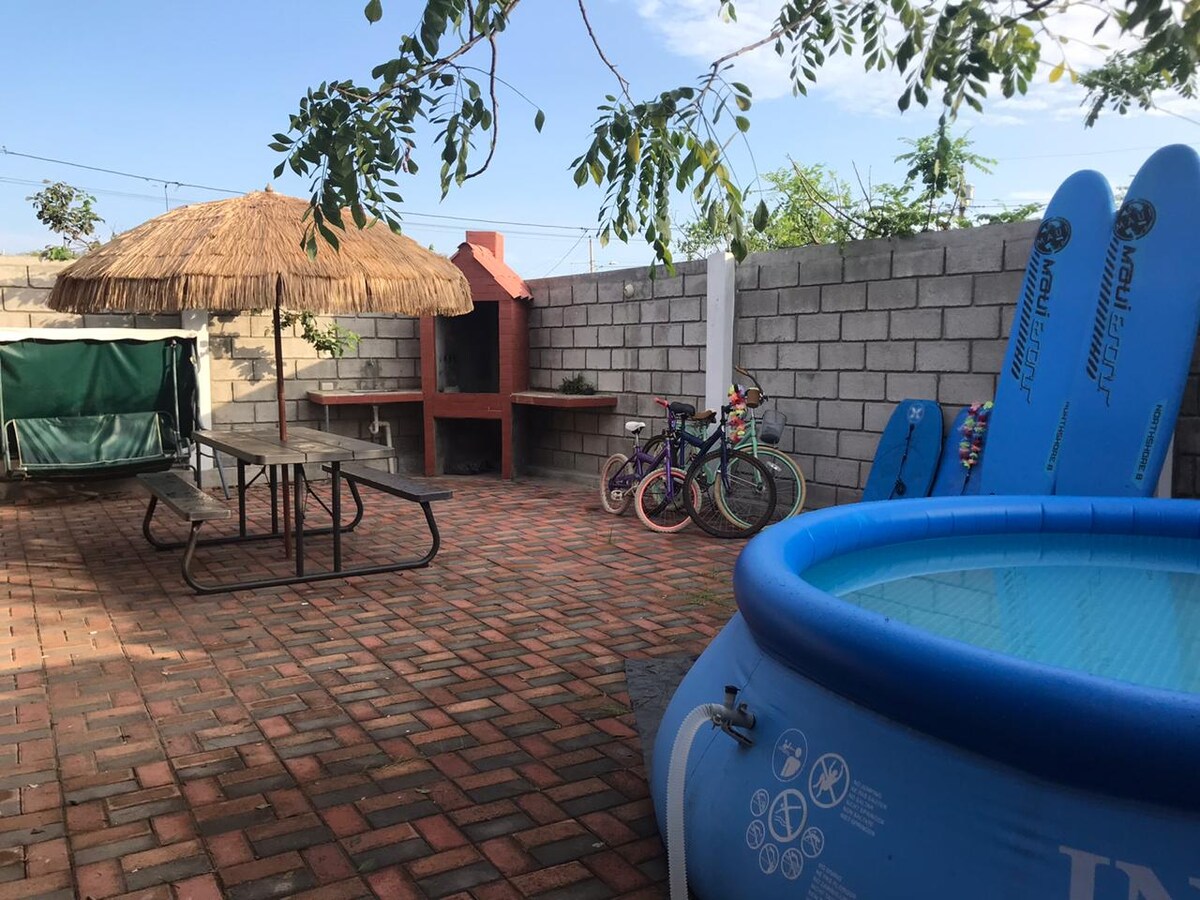 An outdoor space is shown featuring a circular pool filled with water, surrounded by colorful bicycles. A picnic table sits underneath a thatched umbrella, while a barbecue area is visible in the background. Surfboards lean against the wall, enhancing the recreational ambiance.