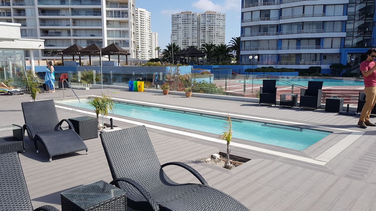 The outdoor pool area is surrounded by modern lounge chairs and features a well-maintained pool. The spacious deck is complemented by palm trees and umbrellas, providing relaxation opportunities. In the background, the building's architecture and surrounding greenery enhance the vibrant atmosphere of the space.
